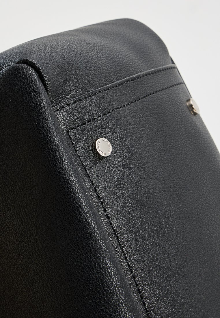 Ecke einer schwarzen, strukturierten Lederhandtasche mit sichtbaren Nähten, silberner Hardware und dezentem Branding auf einer glatten Oberfläche.