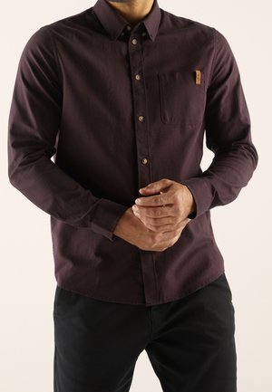 Hombre con camisa de botones color púrpura oscuro con bolsillo en el pecho y pantalones negros, manos entrelazadas al frente sobre fondo liso.