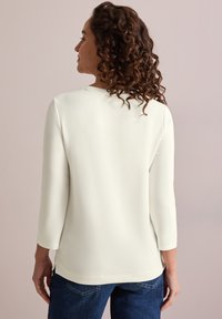 White, mid-length sleeve shirt with a rounded neckline, smooth texture, and side slits at the hem, paired with blue jeans.