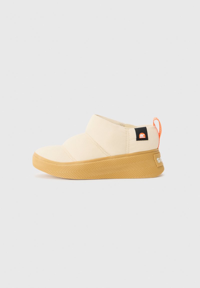 Zapatos slip-on color beige claro con suela de goma, parte superior texturizada y una pestaña de tirado naranja. Presenta una placa de logo negra en el costado.