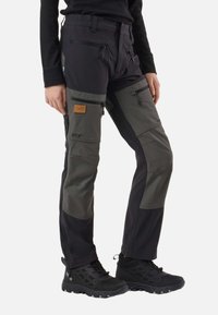 Personne portant un pantalon d’extérieur noir et gris foncé avec plusieurs poches zippées et des chaussures de randonnée noires, debout sur un fond blanc.
