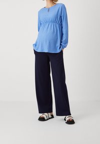 Ljusblå strukturerad långärmad topp med nyckelhålsdetalj, kombinerad med marinblå vida byxor och svarta sandaler med vita accenter.