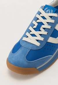 Zili sportiskie apavi, kas izgatavoti no kombinētiem zamšādas un sieta materiāliem, ar baltām auklām un elastīgu gumijas zoli. Ievērojami perforēti izmēri augšdaļā.
