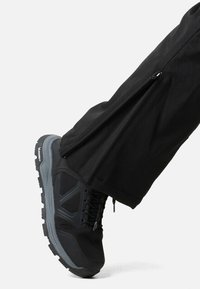 Zwarte wandelschoen met grijze accenten en een gestructureerde zool, gecombineerd met zwarte broek met een zijritsdetail.