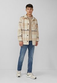 Beige geruite overhemdjasje met donkergrijze strepen, knoopsluiting aan de voorkant, borstzak, gecombineerd met een lichte sweater, blauwe jeans en meerkleurige sneakers.