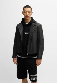 Giacca da motociclista nera in pelle con chiusura a zip e imbottitura sulle spalle, indossata sopra un hoodie nero e pantaloni corti neri con dettagli del logo.
