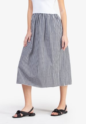 Woman wearing a black and white checkered midi skirt, white top, and black slingback sandals with knotted straps, standing on white background.