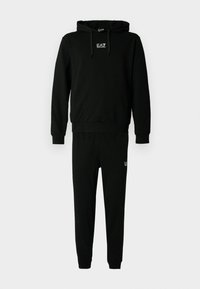 Melnas kapuces džemperis un bikses komplekts ar baltu EA7 Emporio Armani logotipu uz krūtīm un augšstilbiem, attēlots uz baltas fona.