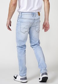 Jeans de denim azul claro con un corte ajustado, que cuentan con bolsillos traseros y un parche de cuero con logo en la cinturilla. Se llevan con zapatillas blancas y negras.