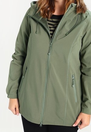 Femme portant une veste à capuche zippée vert clair avec des poches latérales zippées et une chemise rayée en dessous.
