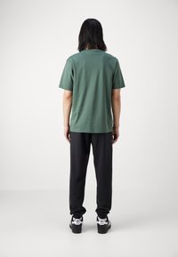 Zelené bavlněné tričko s krátkým rukávem, v kombinaci s černými tepláky. Model nosí černé tenisky s bílým logem. Jednoduchý, pohodlný design.