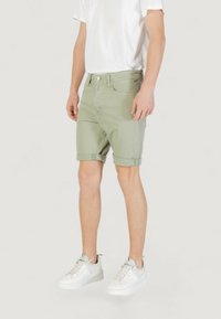 Pantalones cortos de mezclilla verde claro con dobladillos enrollados, bolsillos delanteros y cierre de botón, combinados con una camiseta blanca sencilla y zapatillas blancas.