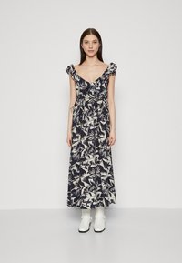 Robe maxi avec un motif floral bleu marine et crème, bretelles froncées, décolleté en V profond, taille cintrée, portée avec des bottines blanches.