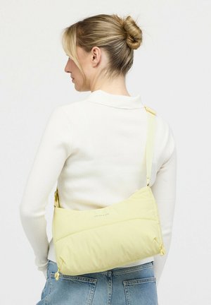 Frau mit blondem Haar im Dutt trägt einen weißen, gerippten Pullover und blaue Jeans und hat eine hellgelbe Umhängetasche auf dem Rücken.