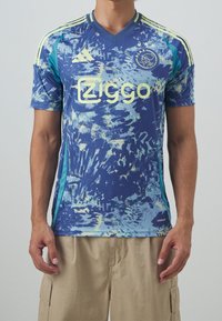 Niebieski i turkusowy t-shirt z krótkim rękawem, z abstrakcyjnymi wzorami, z logo "Ziggo" i herbem Ajaxu, wykonany z lekkiego, oddychającego materiału.