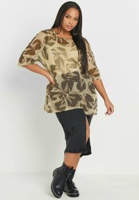 Top beige trasparente con stampa a foglia e maniche ampie, abbinato a una gonna di jeans nera con spacco laterale e stivali neri con lacci.