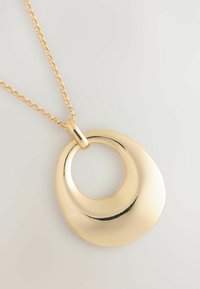 Necklace u zlatnoj boji s okruglim privjeskom koji ima zakrivljen, glatki dizajn. Lanac je delikatan, što upotpunjava polirani završetak privjeska.