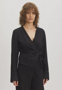 Schwarze Wickelbluse mit langen Ärmeln, Kordelzug-Detail an der Taille und tiefem V-Ausschnitt. Hergestellt aus einem glatten, leichten Stoff mit einem subtilen Fall.