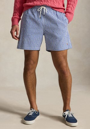 Bărbat purtând pantaloni scurți în carouri alb-albastre cu șnur, pulover roz cu model torsadat și adidași bleumarin din pânză cu șireturi albe.