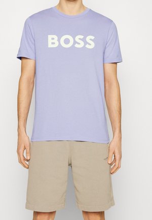 Personne portant un t-shirt violet clair avec "BOSS" en lettres blanches et un short beige, debout devant un fond uni.