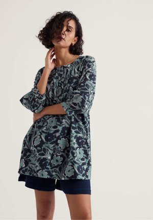 Göndör hajú nő laza, virágmintás tunikában és sötét rövidnadrágban, egyik kezét a nyaka közelében tartva, a másikat a derekán keresztbe téve pózol.