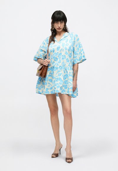 Γυναίκα που φοράει κοντό μπλε και λευκό φόρεμα με λουλούδια και μανίκια μέχρι τον αγκώνα, κρατώντας καφέ τσάντα ώμου και φορώντας σκούρα πέδιλα με ανοιχτά δάχτυλα.