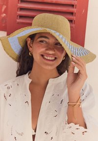 Cappello di paglia a tesa larga in verde oliva chiaro con accenti blu; camicia bianca ricamata con motivi floreali; indossa orecchini a cerchio dorati.