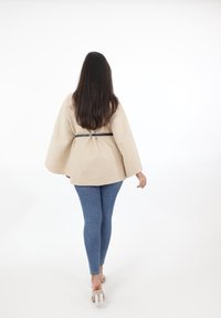 Beige oversized jacka med vida ärmar, åtdragen i midjan med ett svart bälte, tillsammans med blå skinny jeans och klara högklackade skor.