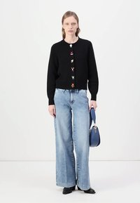 Crna pletena cardigan s šarenim cvjetnim dugmetima, uparena s laganim plavim širokim trapericama i plavom torbom na ramenu. Glatka tekstura.