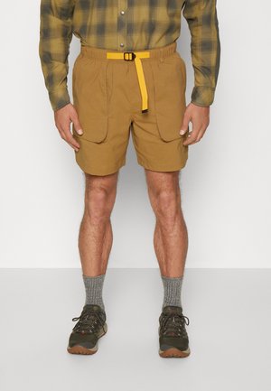 Beige Shorts mit elastischem Bund und gelbem Schnallen-Gürtel. Mit zwei vorderen Taschen. Kombiniert mit grau gestrickten Socken und grünen Schuhen.