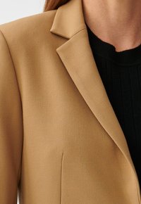 Lichtbruine op maat gemaakte blazer met een gestructureerde stof, met kraagmet inkeping en een enkele borstzak, gedragen over een zwart geribd topje.