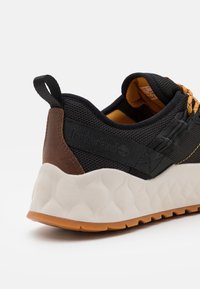 Zapatilla deportiva de malla negra con detalles en cuero marrón, suela texturizada beige y cordones naranjas. Logo en relieve en el lateral y tira para tirar en el talón.