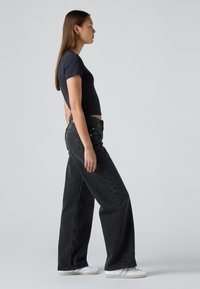 Mujer de perfil que lleva una camiseta negra corta, unos pantalones vaqueros anchos negros y zapatillas blancas, contra un fondo gris liso.