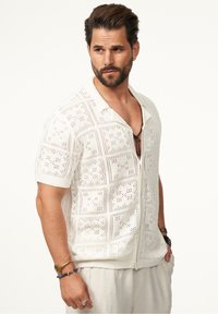 Weißes, kurzärmliges Strickshirt mit offenem Muster, V-Ausschnitt und Reißverschluss auf der Vorderseite. Strukturierten Stoff mit kastigen Löchern und Bündchen.