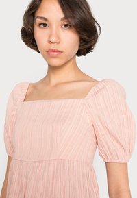 Vestido texturizado rosa claro com decote quadrado e mangas bufantes. Apresenta riscas verticais e um corpete ajustado que se transforma numa saia solta.