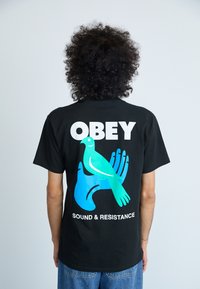 Musthave musta lühikeste varrukatega T-särk, millel on graafiline kujundus: sinine lind käe peal, ning sõnad "OBEY" ja "SOUND & RESISTANCE" valgetes tähestikes.