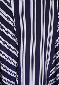 Granatowa bluzka z białymi pionowymi i diagonalnymi paskami. Gładki, lekki materiał o błyszczącym wykończeniu i subtelnym połysku.