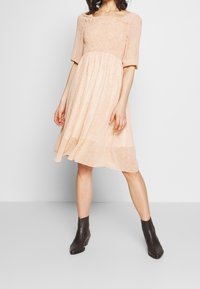 Žena v broskvové šaty ke kolenům s malými květinovými vzory a černými kotníkovými botami, stojící na světlém jednobarevném pozadí.