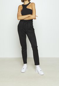 Svarta, högmidjade papperspåsebyxor med knytbälte, avsmalnande vristerna, tillverkade av mjukt, strukturerat tyg, i kombination med en svart ärmlös topp och vita sneakers.