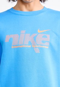 Blå bomuldstrøje med Nike-logoet i prikket orange, med teksten "International Sportswear" nedenfor i fed skrift.