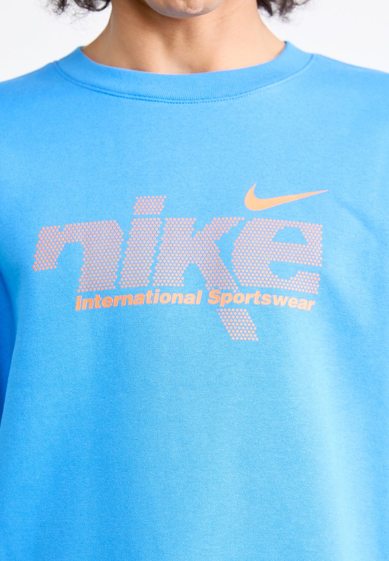 Niebieska bawełniana bluza z kapturem z logo Nike w pomarańczowych kropkach, poniżej z tekstem "International Sportswear" w wyraźnej czcionce.