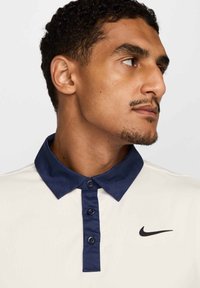 Koszulka polo Nike z kremowym korpusem i granatowym kołnierzykiem, z zapięciem na guziki oraz małym czarnym logo na piersi. Gładka tekstura.