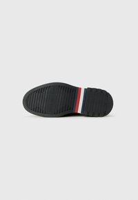 Suela de zapato negra con patrón de tracción acanalado, que presenta franjas rojas, blancas y azules cerca del talón sobre un fondo blanco liso.