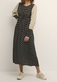 Kvinna iklädd en svart och beige prickig midiklänning med puffärmar och beige sneakers, stående mot en ljus, enfärgad bakgrund.