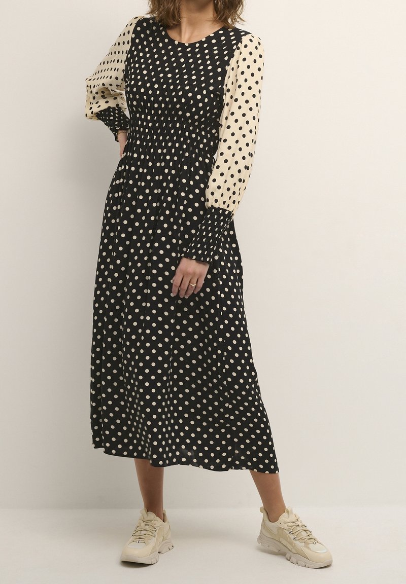 Kvinna iklädd en svart och beige prickig midiklänning med puffärmar och beige sneakers, stående mot en ljus, enfärgad bakgrund.