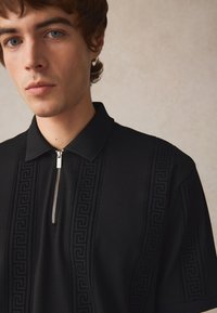 Černé polo tričko s zipovým výstřihem, texturovaným límcem a embosovaným řeckým vzorem na přední části. Hladký povrch látky.