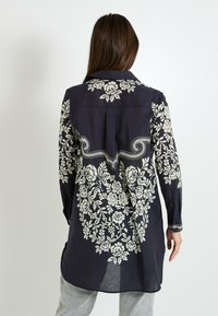 Camisa de manga larga en color azul marino con bordado floral blanco en los hombros y la espalda, con cuello mao y dobladillo curvado.