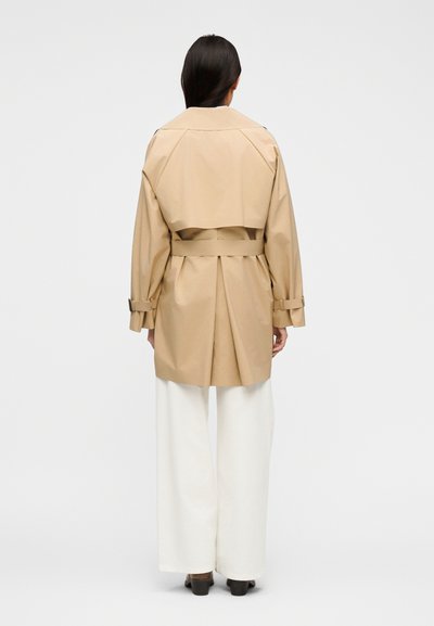 Γυναίκα με μακριά σκουρόχρωμα μαλλιά, ντυμένη με μπεζ παλτό τύπου καμπαρντίνα με ζώνη, λευκό παντελόνι φαρδιάς γραμμής και μαύρα παπούτσια με τακούνι, όρθια με την πλάτη προς τα εμπρός.