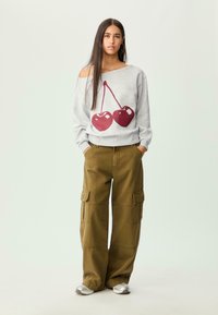 Sweatshirt gris avec un grand graphique de cerise, design décalé sur les épaules, associé à un pantalon cargo olive avec poches latérales et jambes fuselées.