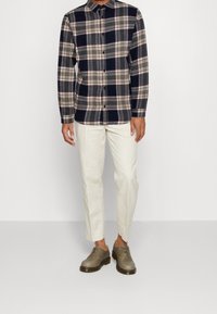 Koszula flanelowa w kratę w kolorach granatowym, kremowym i czerwonym; białe lekkie spodnie; szare sznurowane buty z czarną podeszwą. Widok z przodu, model stojący.